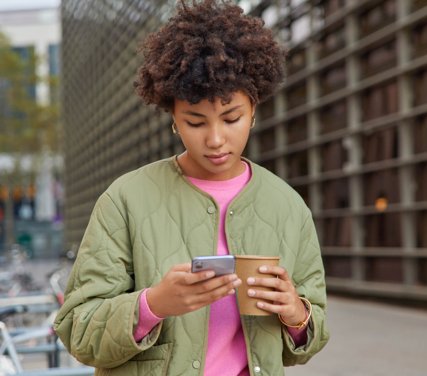 outdoor-shot-curly-haired-young-woman-drinks-aromatic-coffee-surfs-internet-mobile-pone-wears-jacket-watches-video-online-spends-free-time-city-poses-against-blurred-background