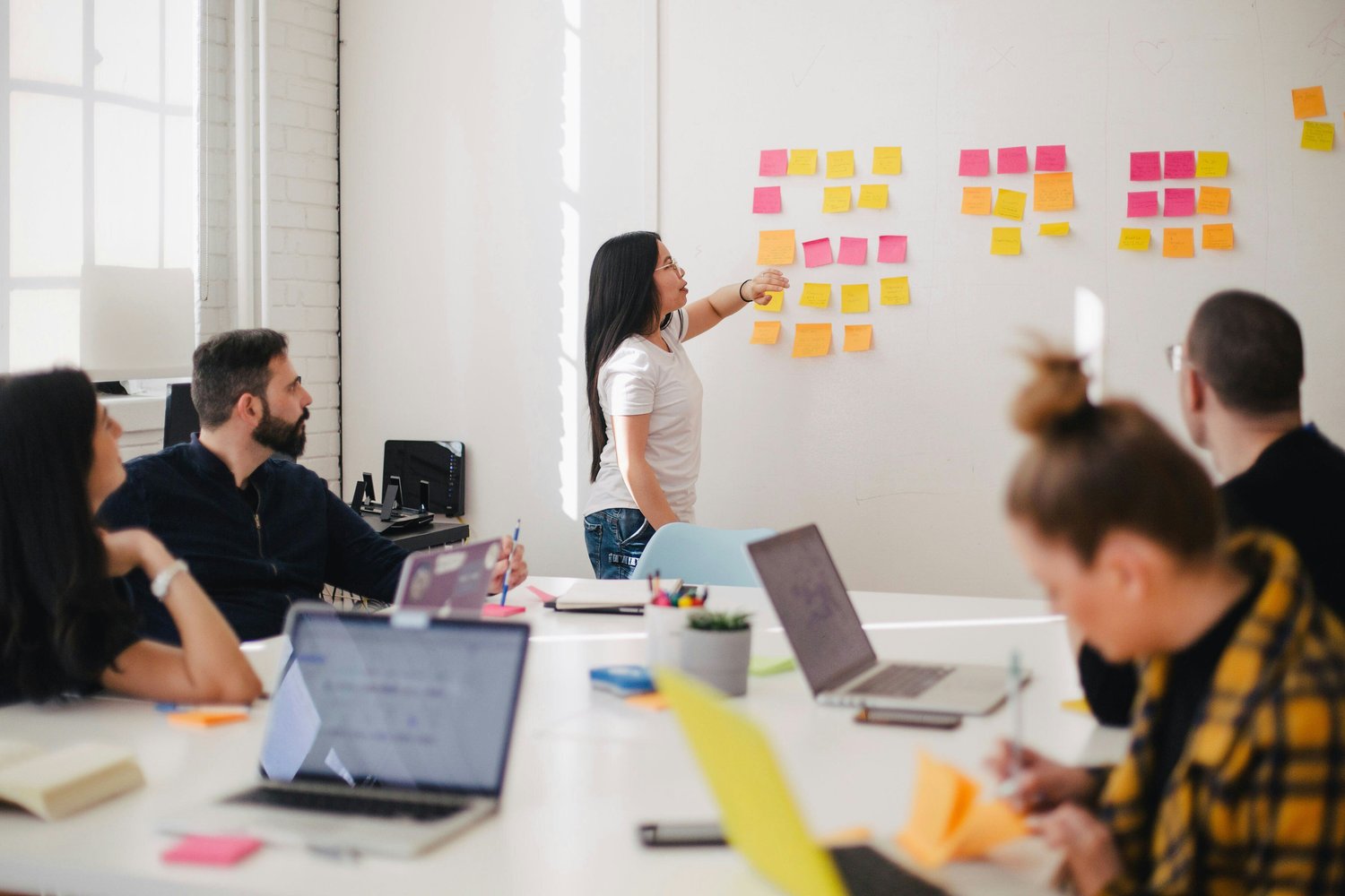 A team of five people are working together in a white meeting room. There is a whiteboard at the back of the room covered in multiple, colorful post-it notes and one person is gesturing to and talking about them. The rest of the team are sat at a large white desk with laptops out, discussing the work with the group or making their own notes. 